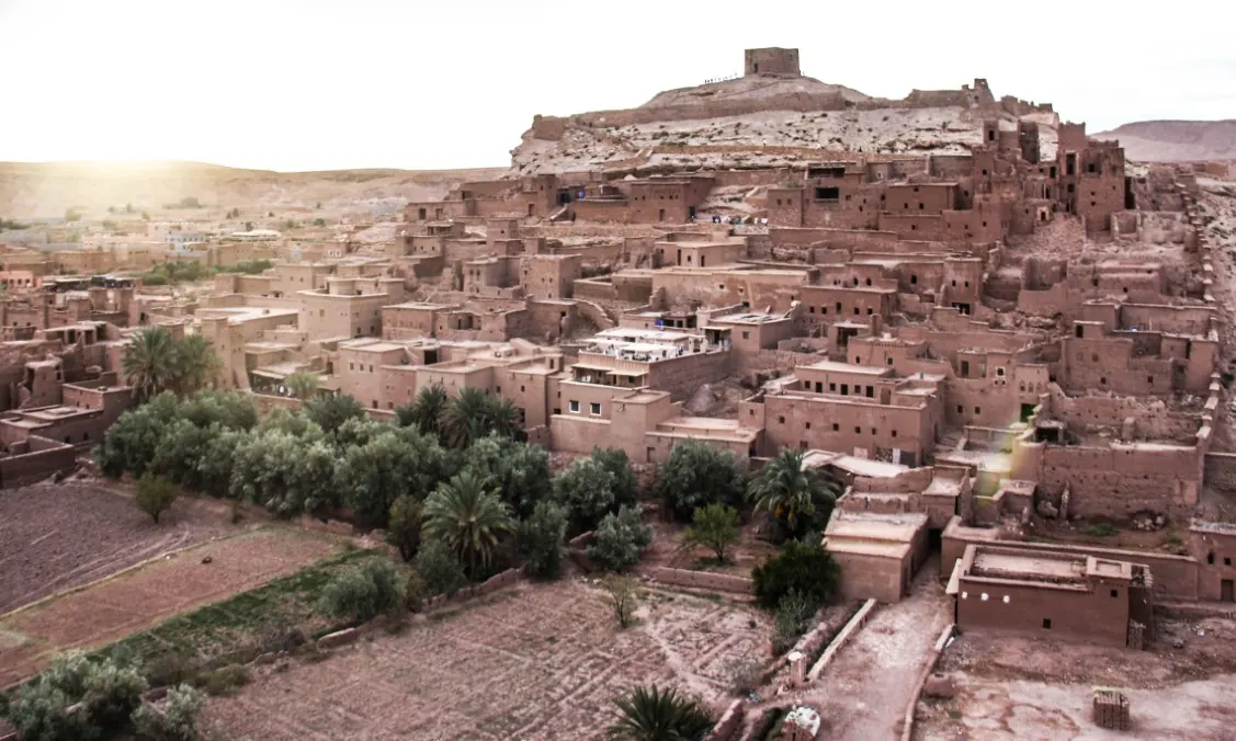Ait Ben Haddou ksar rising up its hillside, with earthen houses, palm trees and fields below at sunset