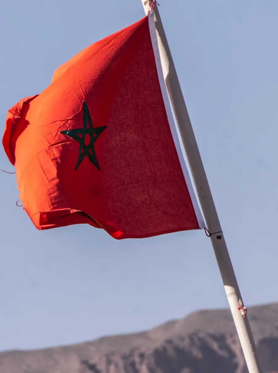 Bandera marroquí en un mástil blanco frente a un cielo claro, con montañas áridas al fondo