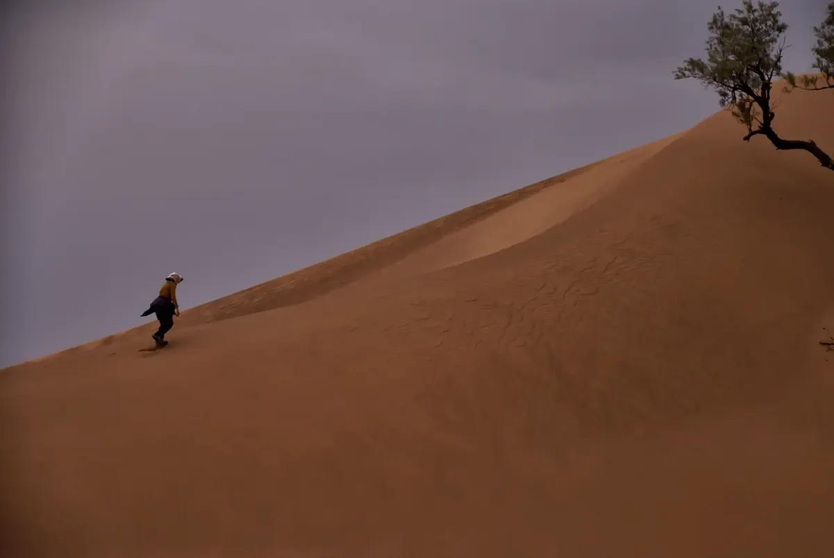 Randonneur montant une grande dune sous un ciel orageux au Maroc