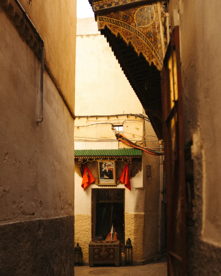 Two Moroccan flags hung on either side of a royal portrait above a doorway in a narrow medina lane