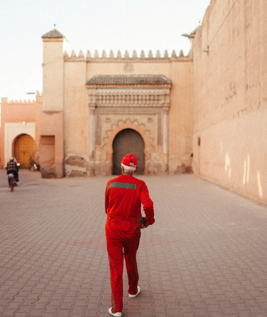 A man in a red tracksuit with MOROCCO written in green on the back, walking through an old town