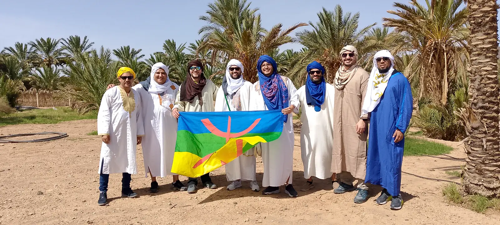 A group of travellers in white djellabas and turbans holding the Amazigh cultural symbol in a palm grove in Morocco