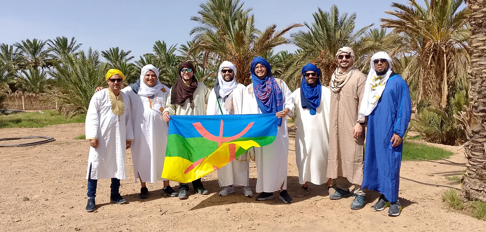Grupo de viajeros con chilabas blancas y turbantes sosteniendo el símbolo cultural amazigh en un palmeral de Marruecos
