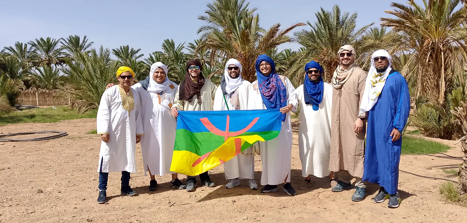 Groupe de voyageurs en djellabas blanches et turbans tenant le symbole culturel amazigh dans une palmeraie au Maroc